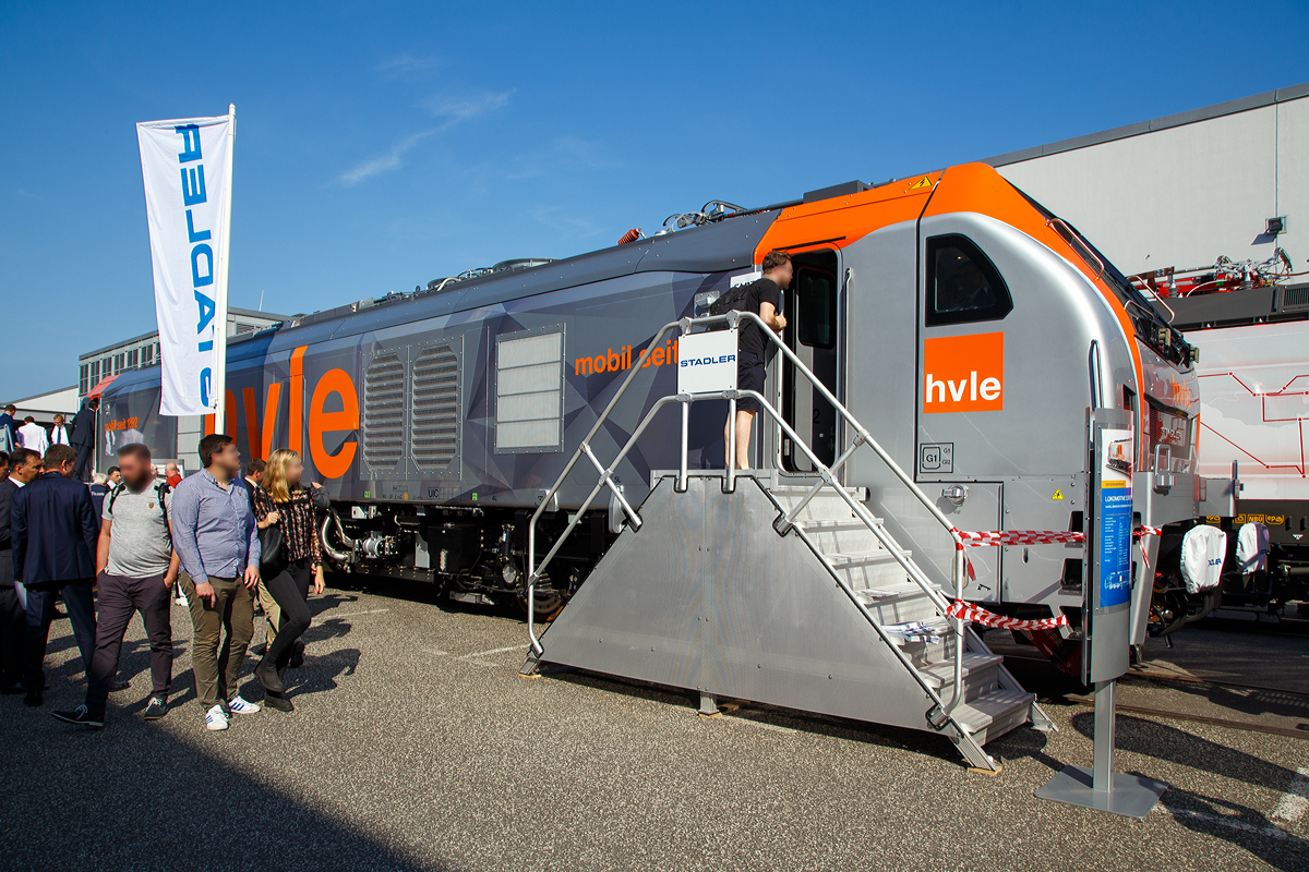 Stadler präsentierte auf der InnoTrans 2018 (hier 19.09.2018) in Berlin seine sechsachsige Eurodual eine Hybrid- bzw. Dual-Mode-Lokomotive. Hier in Form der für die Havelländische Eisenbahn AG (hvle) bestimmte 159 003-3 (90 80 2159 003-3 D-HVLE).
Leider war die ganze Messe so sehr überlaufen, dass ein Foto ohne Menschen davor nicht möglich war.

Die Lok wurde 2018 von Stadler Rail Valencia S.A.U. (Spanien) unter der Fabriknummer 3942 gebaut. Die Auslieferung an die hvle erfolgte 2019.

Bei den EURODUAL-Lokomotiven handelt es sich um kombinierte Diesel- und elektrische Lokomotiven für den Streckendienst im Personenverkehr und Güterverkehr, die von Stadler Rail bei Valencia in Spanien produziert werden. Zum 01. Januar 2016 hatte Stadler Rail das Werk bei Valencia von Vossloh übernommen. Die Dual-Mode-Lokomotive lässt sich dank einem elektrischen und einem dieselelektrischen Antrieb sowohl auf elektrifizierten als auch nicht elektrifizierten Eisenbahnlinien einsetzen. Die Lokomotiven zählen zu den größten einteiligen Lokomotiven auf deutschen Gleisen.

Die Konstruktion der Lokomotiven beruht auf Technik des ehemaligen Vossloh-Werks Valencia. Die Lokomotive setzt sich aus Komponenten der Vossloh Euro 4000 (Rahmen und Drehgestelle), der vierachsigen Diesellokomotive UKLIGHT (Class 68) (Tank, Dieselmotoranlage, Lüfter, Abgasanlage) und der bimodalen, auf der UKLIGHT basierenden Class 88 (Haupttransformator, elektrische Ausrüstung) zusammen. Durch die Kombination dieser Elemente entstand die von der HVLE gewünschte Konfiguration der Baureihe. Mit einer Länge über Puffer von 23.020 mm sind die Lokomotiven eine der längsten einteiligen Lokomotivbauarten eines deutschen Betreibers. Die EURODual-Lokomotive von Stadler ist eine Lokomotive mit einer beeindruckenden Leistung, sowohl im Diesel- als auch im Elektroantriebsmodus.

Die tragenden Elemente der Lok sind aus verschiedenen Stahlsorten hergestellt. Die Seitenwände des Maschinenraumes sind fest mit dem Rahmen verschweißt. Führerhäuser und Einstiegsräume sind separate Aufbauten. Das Dach besteht aus fünf Segmenten, sie nehmen neben den Stromabnehmern auch andere Komponenten wie die Kühlerlüfter auf. Die Führerstände sind schallisoliert und klimatisiert. Der Lokführer hat seinen Arbeitsplatz in der Mitte des Führerraumes.

Die Drehgestelle gelten trotz ihrer dreiachsigen Ausführung als gleisschonend, da sie beim Bogenlauf noch geringere Querkräfte als die zweiachsigen Drehgestellen der Bombardier TRAXX und Siemens Vectron aufweisen. Die Primärfederung erfolgt über Schraubenfedern, die Sekundärfederung wird in Form von Gummi-Metall-Elementen und Schwingungsdämpfern realisiert.

Der dieselelektrische Antrieb geschieht durch einen Caterpillar V-16-Viertakt-Dieselmotor mit Abgasturbolader und Ladeluftkühlung vom Typ CAT C175-16 von mit einer Leistung von 2.800 kW, der elektrische Antrieb hat eine Leistung von 6.150 kW bei Speisespannungen von 25 kV / 50 Hz und 15 kV / 16,7 Hz. Die Loks haben Wechselstrom-Antriebssystem mit IGBT. 1 Wechselrichter pro Achse und besitzen eine hohe Anfahr- und Dauerzugkraft.

Die Havelländische Eisenbahn AG (HVLE) ist der erste Kunde, an den die sechsachsigen Lokomotiven mit bimodalem Antrieb, die von Stadler für den europäischen Markt entwickelt und ausgeliefert wurden. Die Lokomotiven der Havelländische Eisenbahn sollen mit unterschiedlichen Fahrdrahtspannungen elektrifizierte und nichtelektrifizierte Strecken bedienen. Konkret betrifft es die mit 25 kV bei 50 Hz elektrifizierte Rübelandbahn, das mit 15 kV bei 16,7 Hz versorgte deutsche Bahnnetz und die bislang nicht elektrifizierten Zulaufstrecken zur Rübelandbahn. Bisher wurde dieser Verkehr elektrisch mit Lokomotiven der Reihe 185 und auf den nicht elektrifizierten Abschnitten mit Diesellokomotiven abgewickelt. Die Eurodual wäre die Universallokomotive für diese Dienste. Außerdem ist sie bedeutend leistungsstärker als die gegenwärtig eingesetzten Maschinen und besitzt dazu noch die für die Steilstrecken notwendige Lokreibungslast. Nach der DR-Baureihe E 251 kommen damit wieder sechsachsige Elektro-Lokomotiven auf der Rübelandbahn zum Einsatz.

Die Zulassung der Lokomotiven für die HVLE erfolgte am 12. Februar 2020. Am 29. August 2020 wurde die (hier gezeigte)159 003 von DB Fernverkehr für Testfahrten auf der Schnellfahrstrecke Köln/Rhein-Main angemietet. Der Testzug bestand aus zwei Triebwagen der Baureihe 407 sowie einer Lokomotive der BR 101. Diese sollte allerdings nur die Masse der Fahrgäste simulieren.

TECHNISCHE DATEN, BR 159 der hlve:
Inbetriebsetzung: 2020
Spurweite: 1.435 mm (Normalspur)
Achsanordnung: Co’Co’
Länge über Puffer: 23.020 mm
Breite: 2.900 mm
Höhe: 4.290 mm
Treibdurchmesser: 1.067 mm (neu)
Eigengewicht: 123 t
Achslast: 21 t
Höchstgeschwindigkeit: 120 km/h
Elektrische Leistung am Rad bei 25 kV 50 Hz: 7.000 kW
Elektrische Leistung am Rad bei 15 kV 16.7 Hz: 6.150 kW
Leistung bei Dieselantrieb: 2.800 kW (3.807 PS)
Anfahrzugkraft: 500 kN
Elektrische Bremskraft : 240 kN

Dieselmotor:
Motorbauart: Caterpillar V-16-Viertakt-Dieselmotor mit Abgasturbolader und Ladeluftkühlung vom Typ CAT C175-16
Motorleistung: 2.800 kW (3.807 PS)
Motornenndrehzahl: 1.800 U/min
Hubraum: 84,67 l (Bohrung Ø 175 mm / Hub 220 mm)
Motorgewicht (trocken): ca. 13.000 kg
Motorabmessung: 4.515 x 1.845 x 2.478 mm (l x b x h)
Abgasnorm: IIIB

Anzahl der Fahrmotoren: 6
Antrieb: elektrisch oder dieselelektrisch
Speisespannung: 25 kV 50 Hz und 15 kV 16.7 Hz
Tankinhalt Diesel: 3.500 l
Tankinhalt Urea: 400 l (für Abgasbehandlung selektive katalytische Reduktion („AdBlue“))
Bremse: KE-GP-E mZ (D)
Bremssystem Mechanisch: Pneumatisch
Bremssystem Dynamisch: Regenerativ/Rheostatisch
Primär Federung: Schraubenfedern
Sekundär Federung: Gummi-Metall Vertikal- und Horizontaldämpfer
Zugbeeinflussung: PZB, ETCS

Bei einer EuroDual die unter 1,5 kV DC Gleichstromoberleitung fährt liegt die 4.100 kW, für eine Leistungssteigerung auf 6.000 kW ist dann auch der Dieselmotor  (im Gleichstrombetrieb) zuschaltbar.

Quelle: Stadler Rail, CAT und Wikipedia 