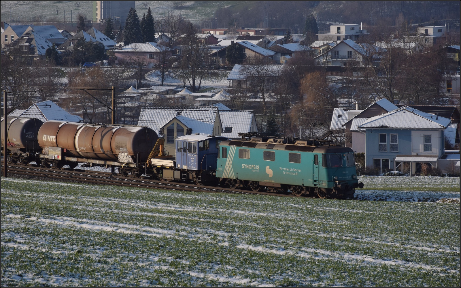 Re 430 114 von WRS mit Synopsis-Werbung schleppt die Rangierhilfe Tm III 9530 mit über den Bözberg. Frick, Dezember 2022.
