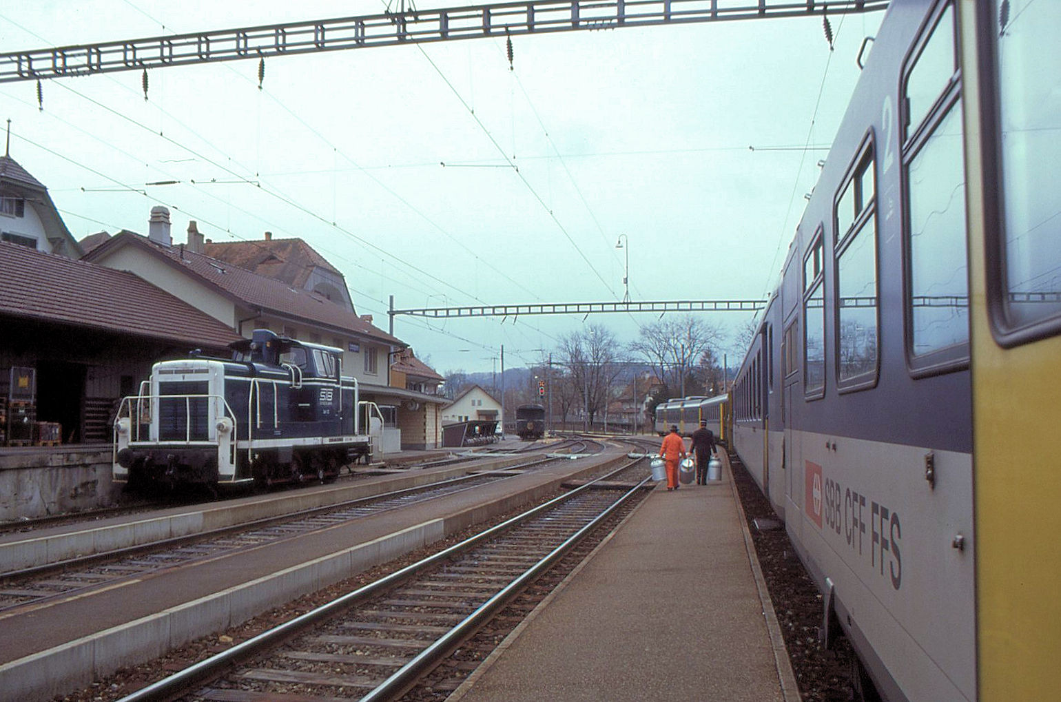 Während im zuvor eingestellten ähnlichen Bild in der Station Laupen der Zugchef des SBB NPZ 2180 Milchkannen herumträgt, erscheint jetzt noch ein oranger Mann mit weiteren Milchkannen... Daneben die ehemals deutsche Em3/3 12 der Sensetalbahn. 4.Januar 1991 
